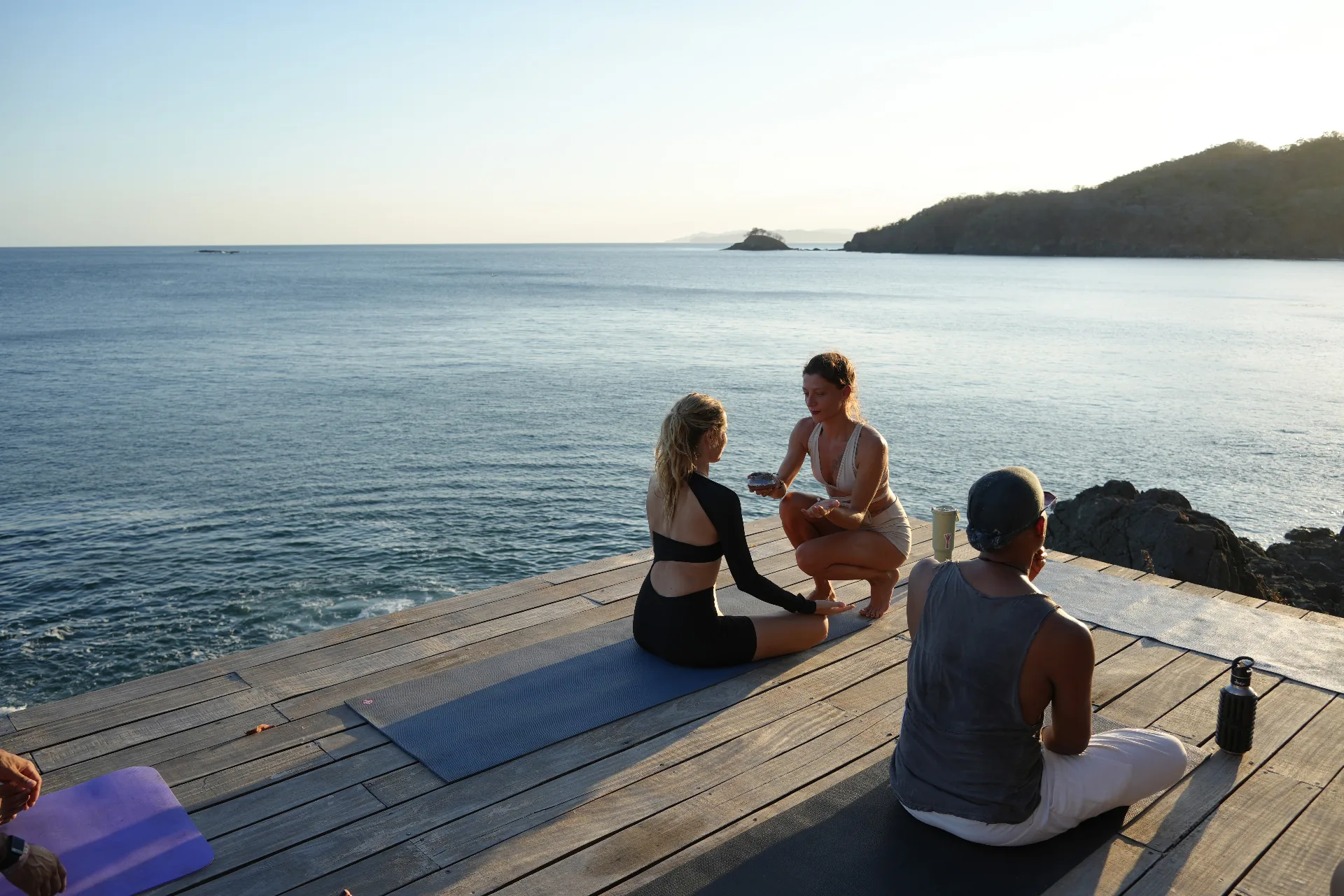 Three people on a wooden deck above the ocean at sunrise, in quiet conversation